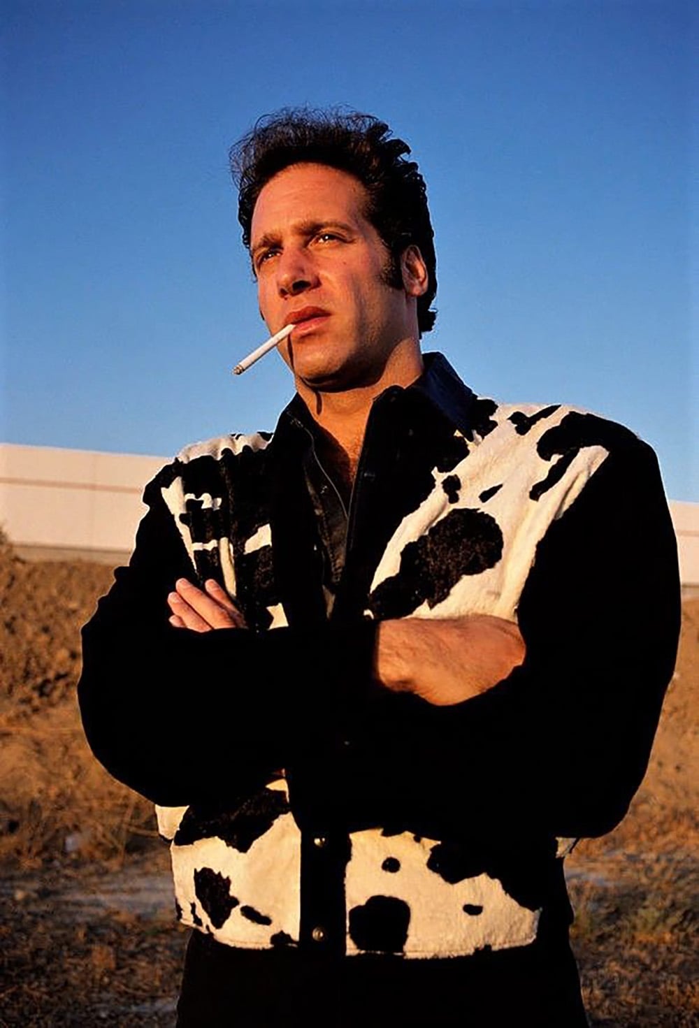 A man stands outdoors with his arms crossed, wearing a black and white cow-print jacket and holding a cigarette in his mouth. The sky behind him is clear and blue, and the lighting suggests late afternoon or early evening.