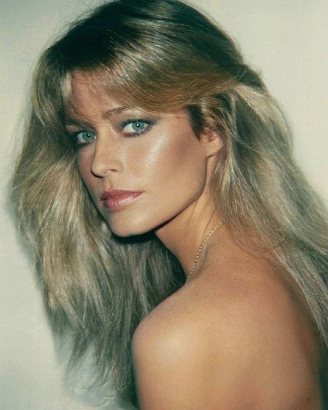 A woman with long, voluminous blonde hair looks over her shoulder at the camera. She has blue eyes, subtle makeup, and is posed against a plain background, with her bare shoulder visible.