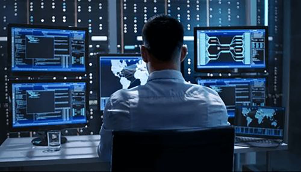A person sits at a desk facing multiple monitors displaying maps and data, in a dark room with servers and blue lights, suggesting a cybersecurity or control center environment.