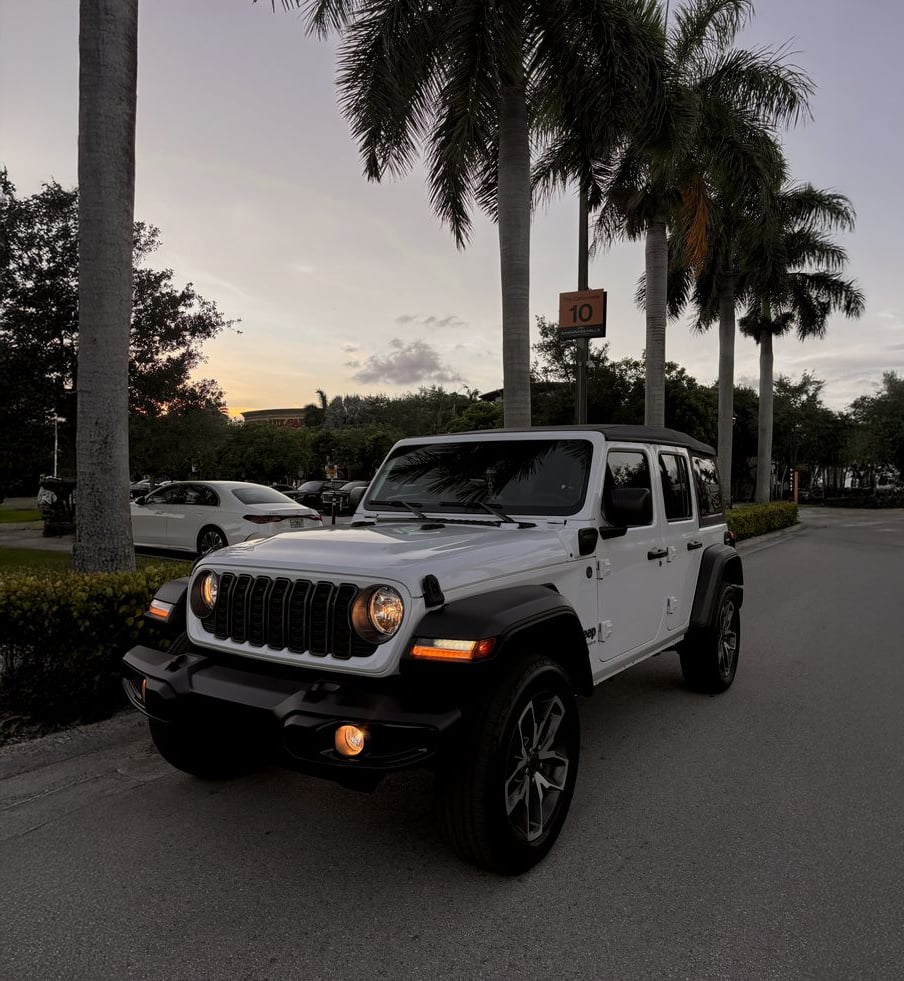 A white Jeep Wrangler with headlights on is parked on a street lined with palm trees at sunset, with a speed limit sign visible and other cars in the background.