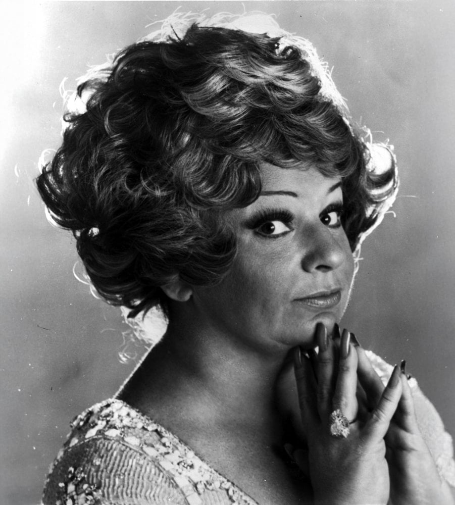 Black and white portrait of a person with wavy, short hair, wearing a sparkly top, dramatic eye makeup, and a large ring, looking at the camera with hands clasped near their face in a thoughtful pose.