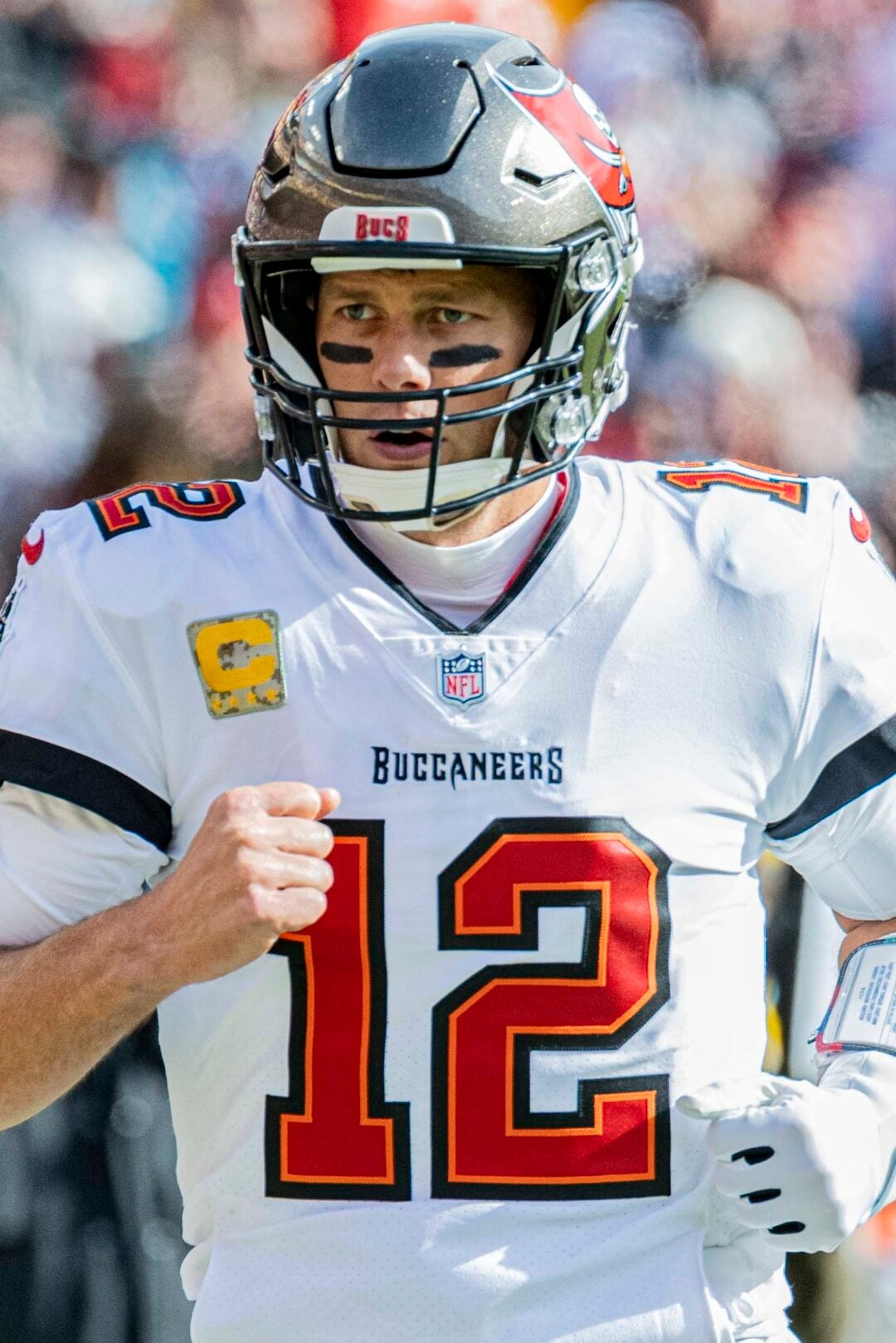 A football player in a white Tampa Bay Buccaneers uniform with the number 12, wearing a helmet and black eye paint, stands on the field during a game.