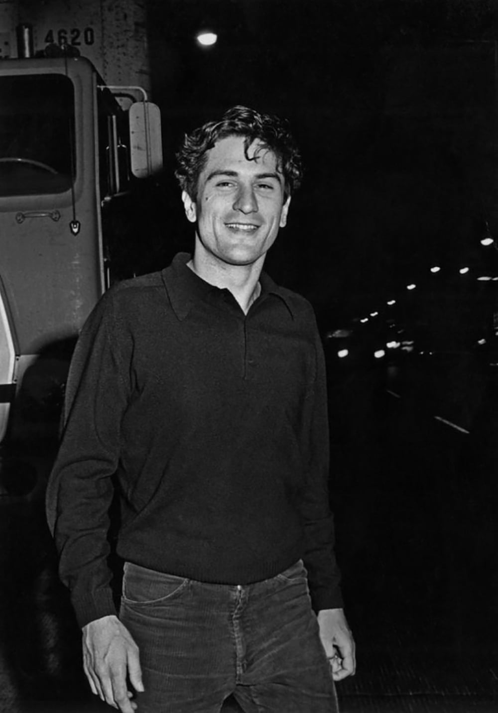 A young man with curly hair, wearing a dark long-sleeve shirt and jeans, stands smiling on a city street at night with streetlights and a large truck in the background.