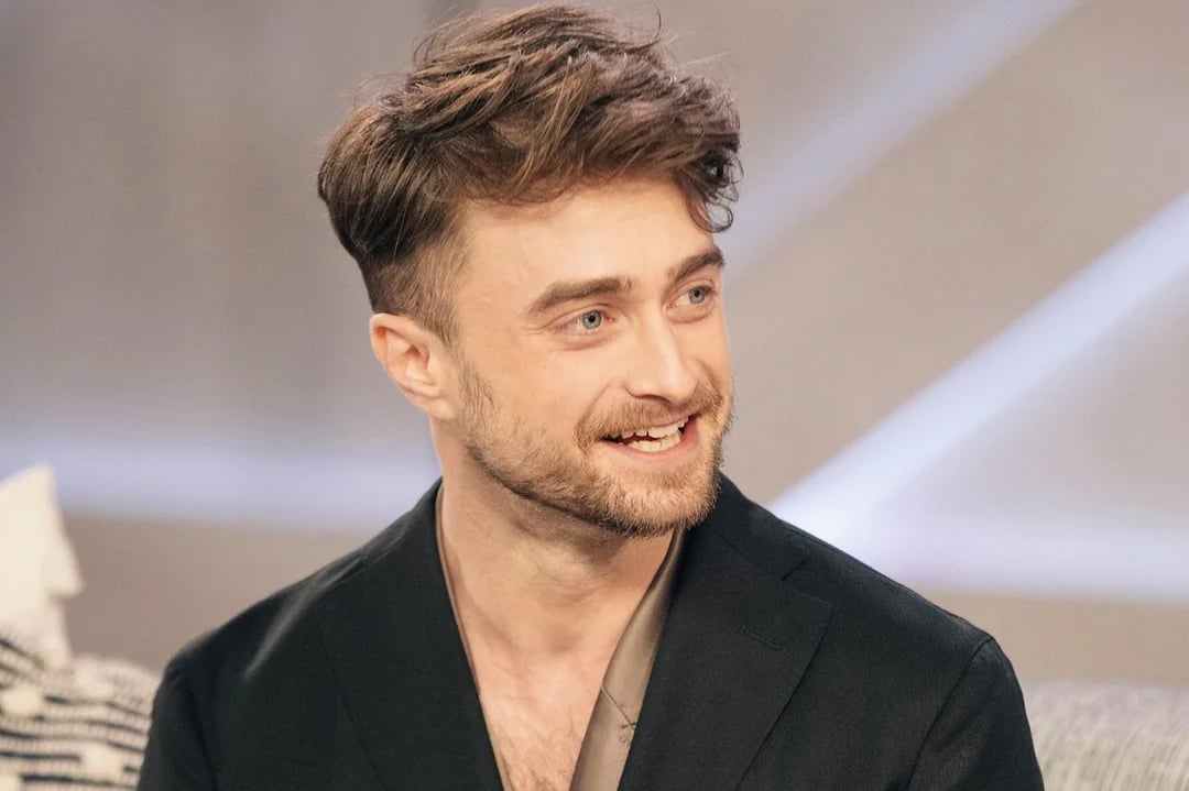 A man with short brown hair and a beard is smiling while looking to the side. He is wearing a dark jacket over a light shirt and is seated in a softly lit indoor setting.