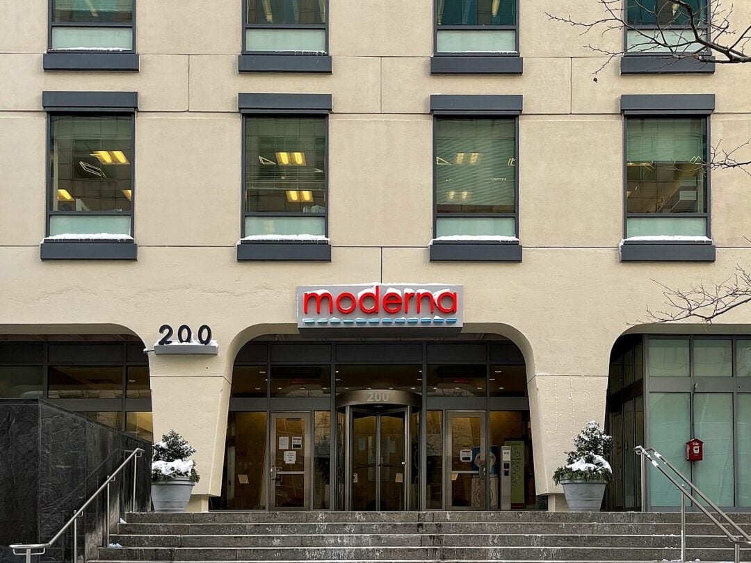 The entrance to a modern office building features a "moderna" sign above glass doors, with "200" displayed on the wall and steps leading up to the entry. Two potted plants sit on either side of the doorway.