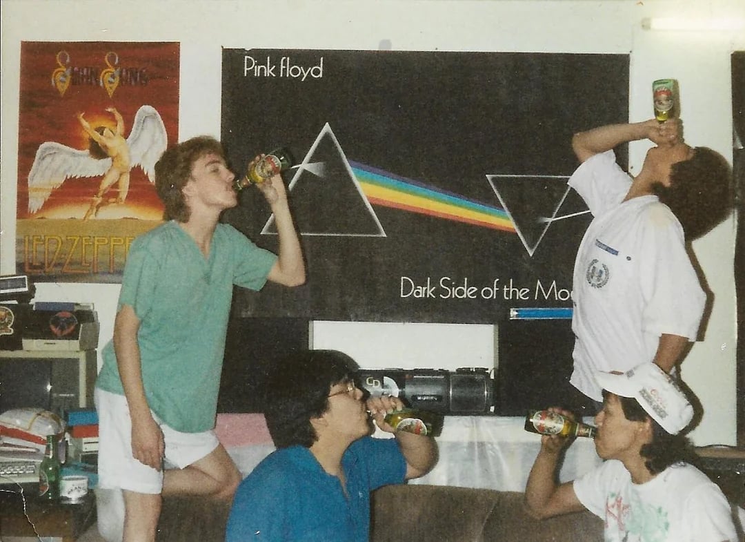 Four people are drinking from cans in a room decorated with Led Zeppelin and Pink Floyd posters. Two are standing, tilting their heads back, while two are seated on a couch, facing each other and drinking.