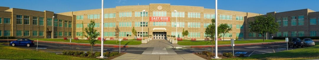 A wide view of a large, three-story school building with many windows, a clock at the top center, a red banner reading “East High School,” and a few cars parked in front on a sunny day.