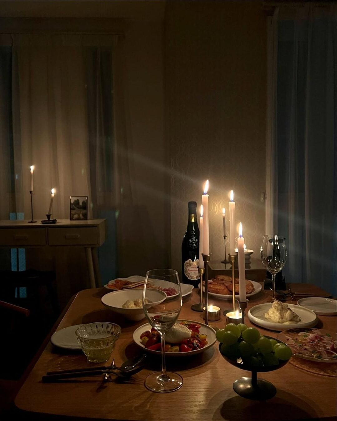 A dimly lit dinner table set for two with wine, lit candles, plates of food, cheese, grapes, and glasses. Soft light creates a cozy, intimate atmosphere. A side table with more candles and a photo is in the background.