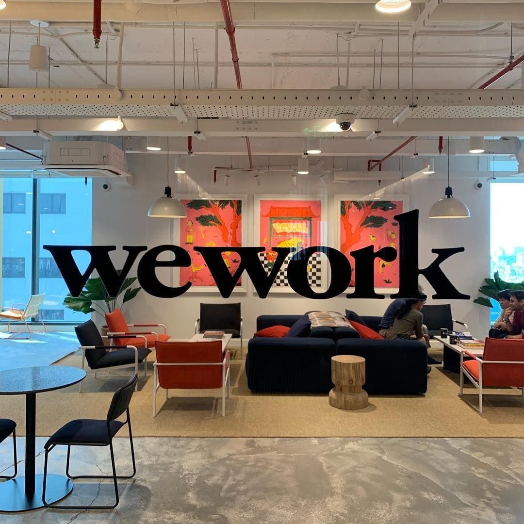 A modern office lounge with “wework” in bold black letters on glass, featuring sofas, tables, potted plants, and colorful artwork on the walls. Several people are sitting and talking. Bright lighting and large windows.