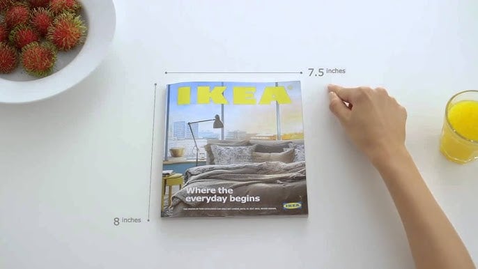 A hand touches an IKEA catalog on a white table, measuring 7.5 by 8 inches. A bowl of rambutan fruit and a glass of orange juice sit nearby. The catalog cover shows a bedroom scene and the text “Where the everyday begins.”