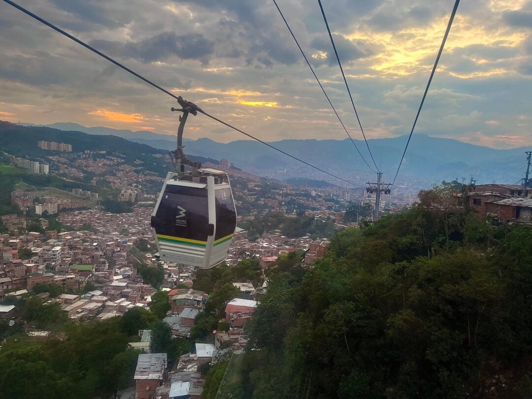 A cable car travels over a densely built urban area with many houses, surrounded by green hills, during sunset with dramatic clouds and mountains in the background.