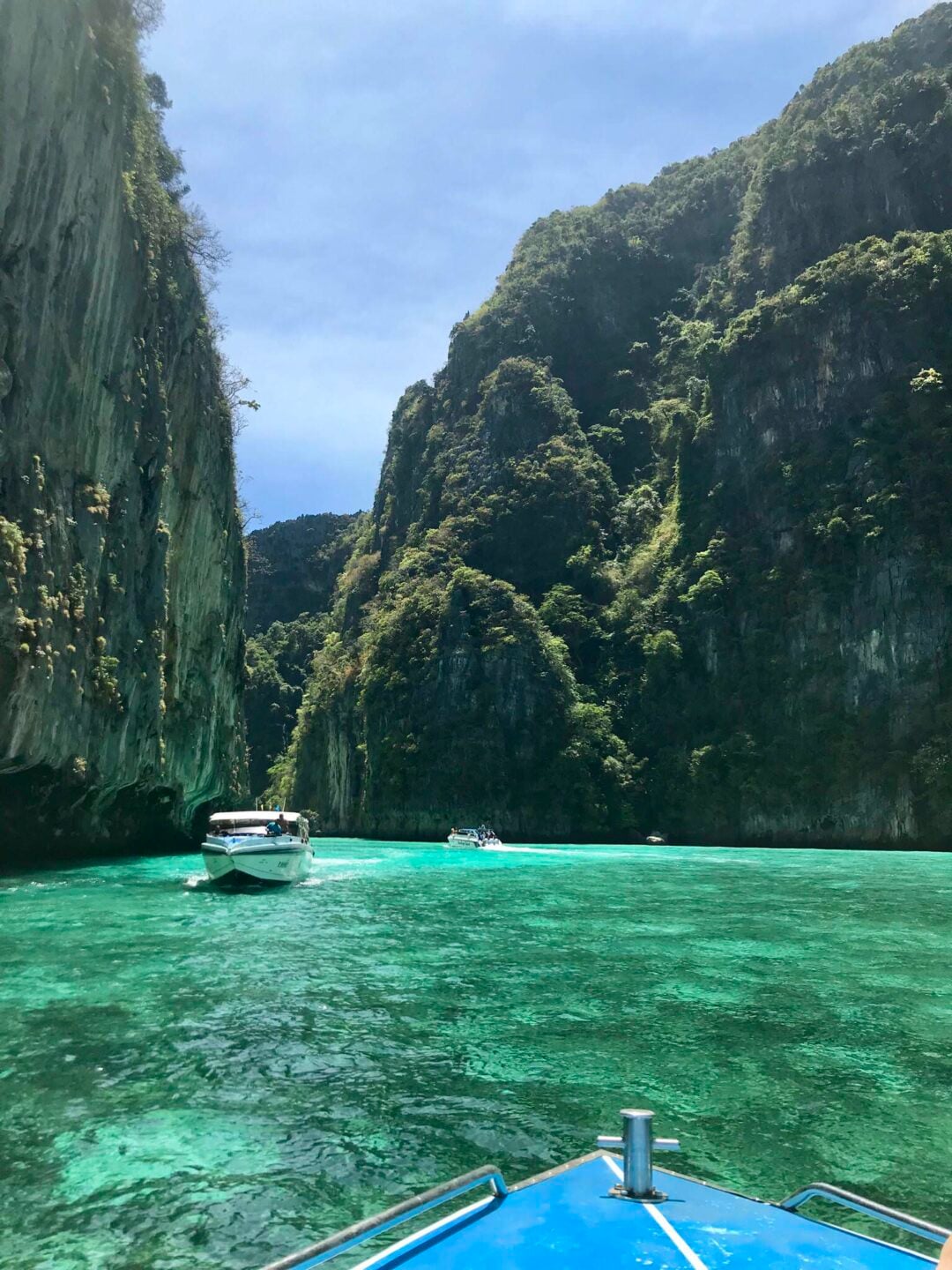 A view from a boat approaching turquoise waters surrounded by tall, lush green cliffs with other boats visible in the clear, sunny lagoon.