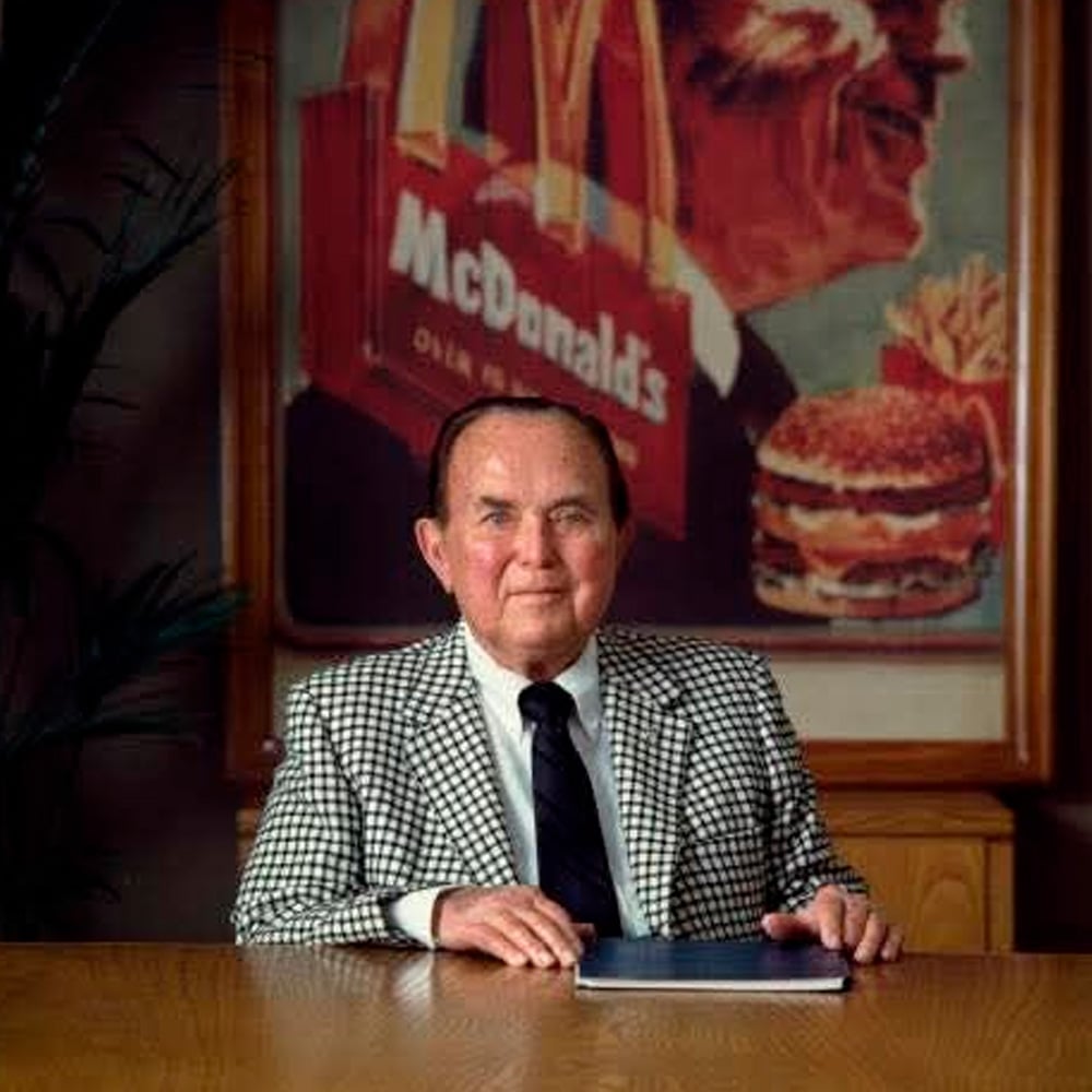 An older man in a checkered suit sits at a wooden table, hand resting on a folder. Behind him is a large artwork featuring McDonald's branding, including a burger, fries, and logo.