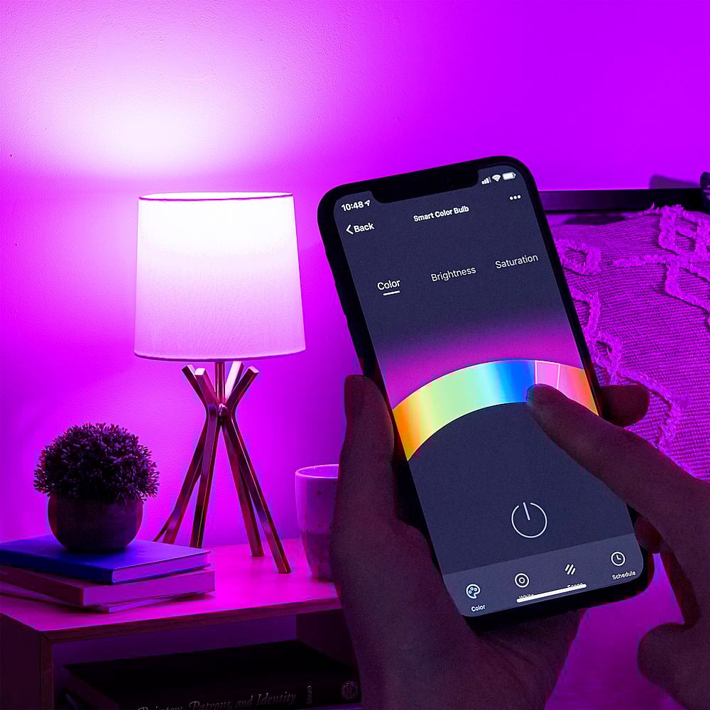 A hand holds a smartphone that controls a lamp’s color, brightness, and saturation. The lamp on a bedside table glows bright pink, casting a purple light across the room. A plant and a cup are also on the table.