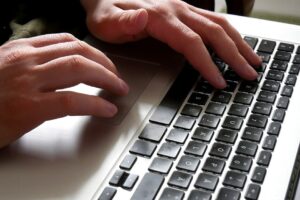 Close-up of a person's hands typing on a laptop keyboard, with one hand on the touchpad and the other hand on the keys.