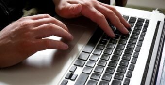 Close-up of a person's hands typing on a laptop keyboard, with one hand on the touchpad and the other hand on the keys.