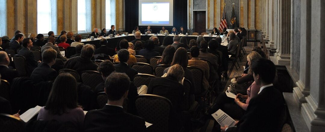 A large group of people attends a formal meeting in an ornate, high-ceilinged room with chandeliers. Panelists sit at a long table facing the audience. A screen displays a presentation at the front. American flags are visible.
