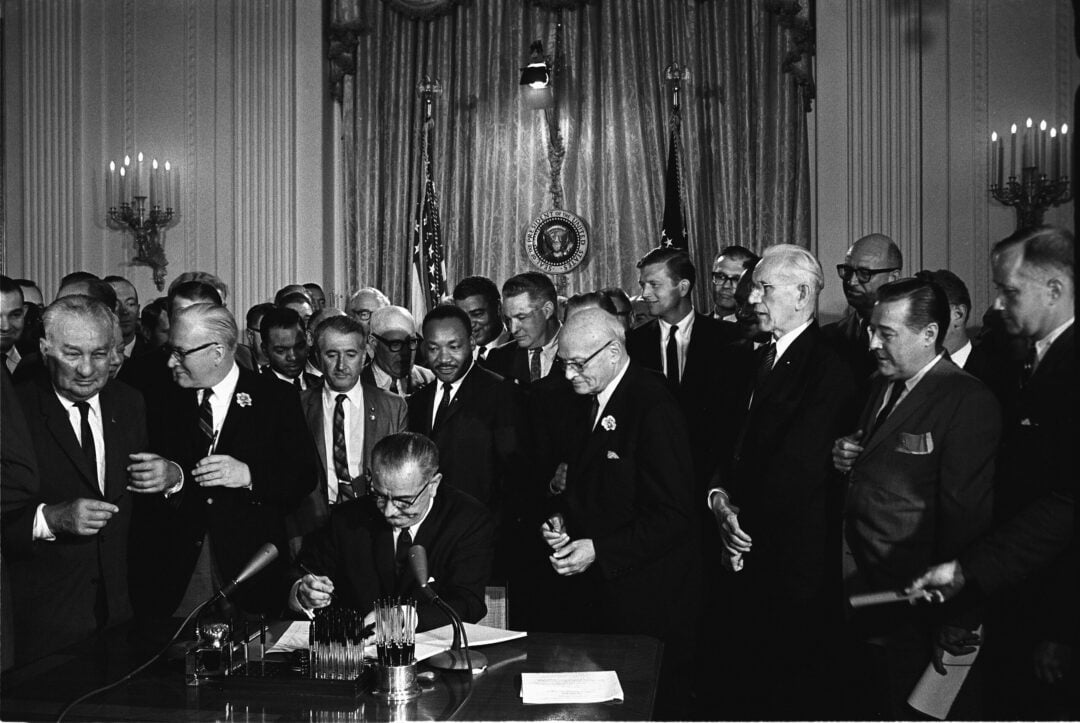 President Lyndon B. Johnson signs the Civil Rights Act of 1964 at a desk, surrounded by a large group of officials and supporters in a formal room with U.S. flags and curtains in the background.
