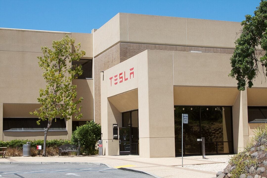 A beige, modern office building with the red Tesla logo above the entrance. There are trees, bushes, and a bench outside, and the sky is clear and blue.