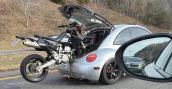 A silver Volkswagen Beetle with its rear hatch open is transporting a motorcycle partially inside the car while driving on a highway. The photo is taken from a nearby vehicle&rsquo;s window, showing other cars in traffic.