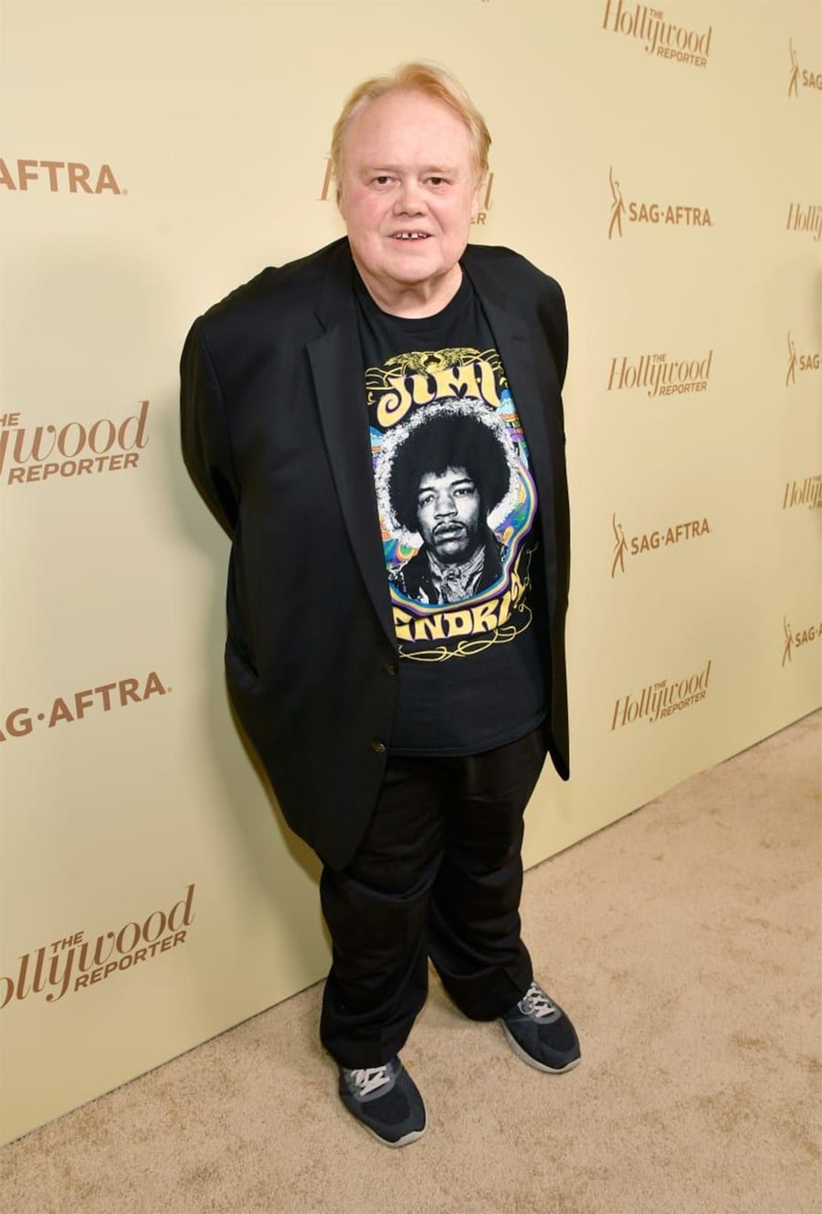 A man with light skin and blond hair stands on a beige carpet, smiling at the camera. He wears a black jacket, black pants, blue sneakers, and a Jimi Hendrix T-shirt. The background features SAG-AFTRA and The Hollywood Reporter logos.