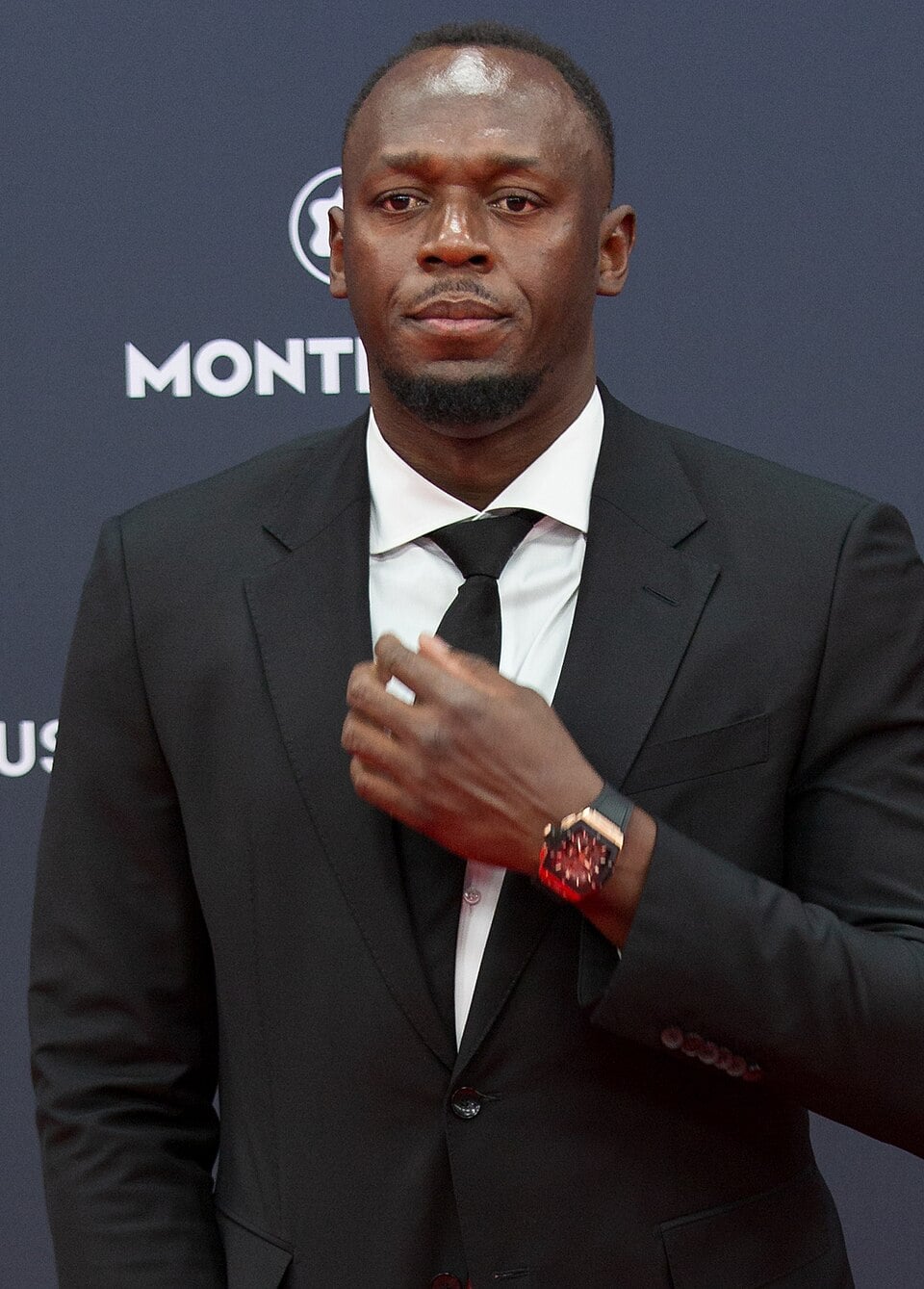 A man in a black suit and tie stands against a dark backdrop, adjusting his tie with one hand. He has short hair and wears a red watch. The background features white logos and text.