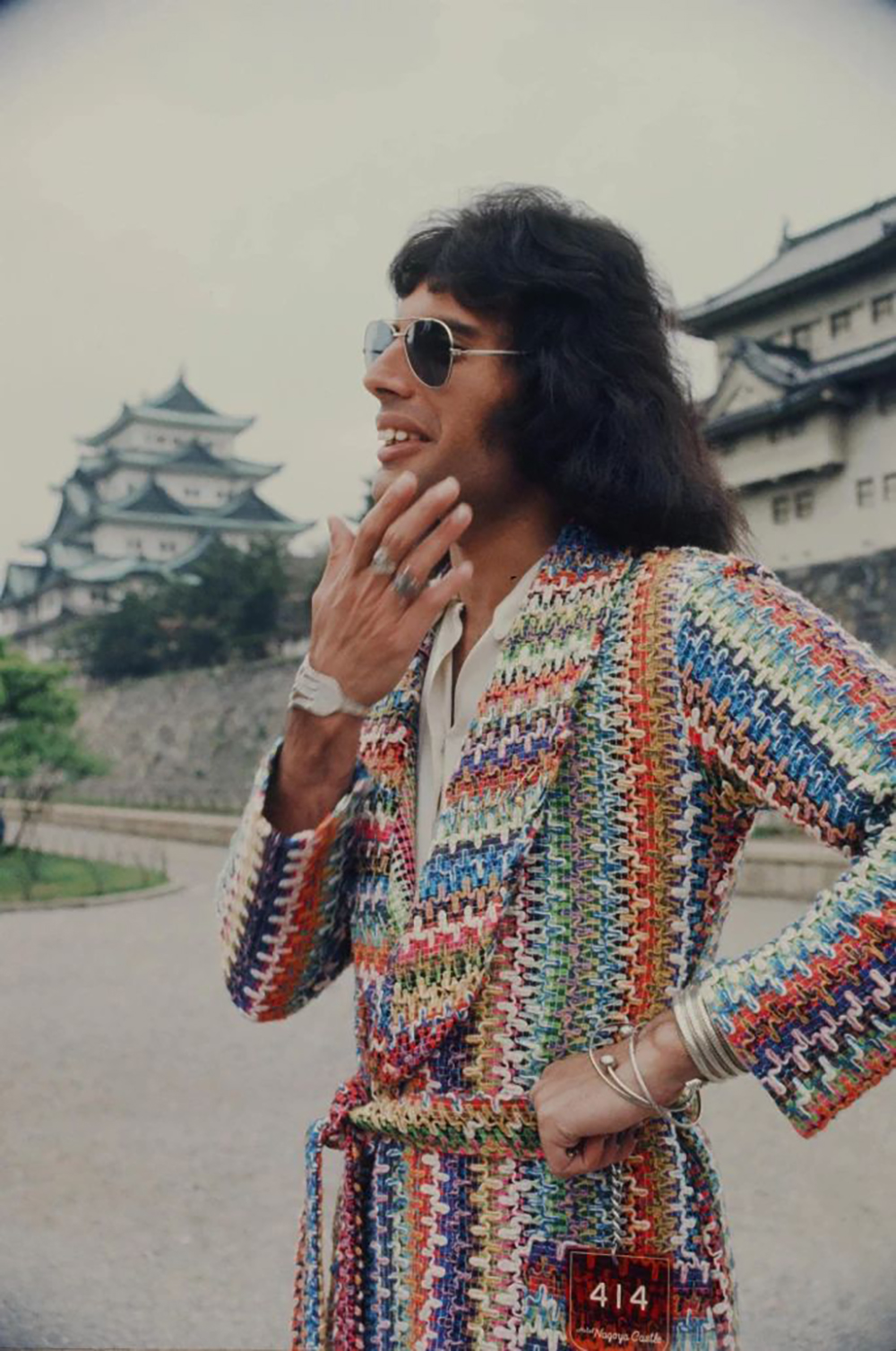 A man with shoulder-length dark hair and sunglasses wears a colorful, patterned jacket and rings, standing outdoors near a traditional Japanese castle.