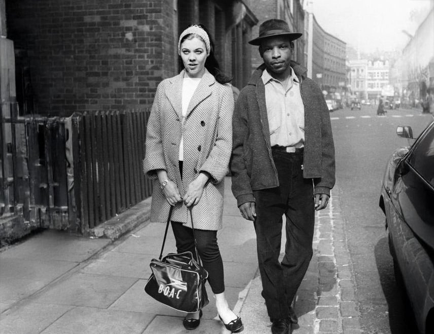 A woman and a man walk side by side on a city sidewalk. The woman wears a headband, coat, and carries a handbag labeled "BOAC." The man wears a hat and jacket. Brick buildings and parked cars line the street.