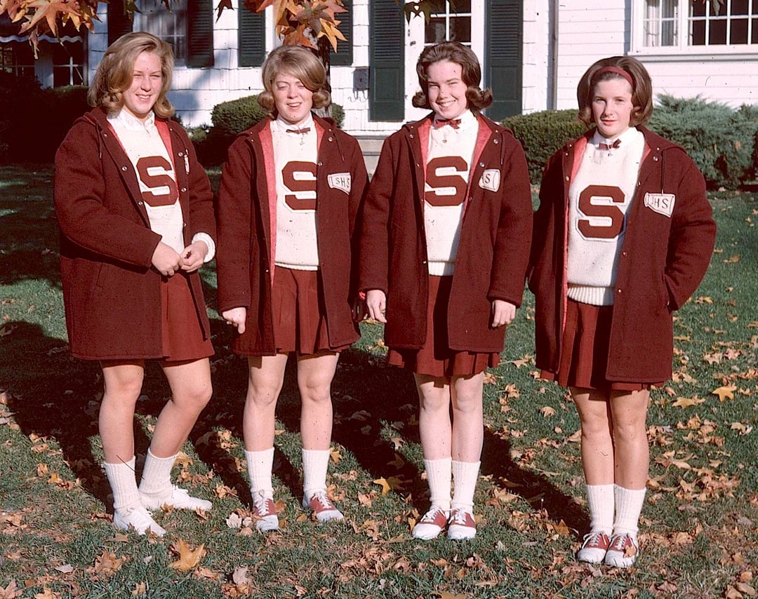 Four teenage girls stand outside on grass, wearing matching maroon and white cheerleading outfits with large "S" emblems, cardigans, skirts, and saddle shoes. Autumn leaves hang above them, and a white house is in the background.
