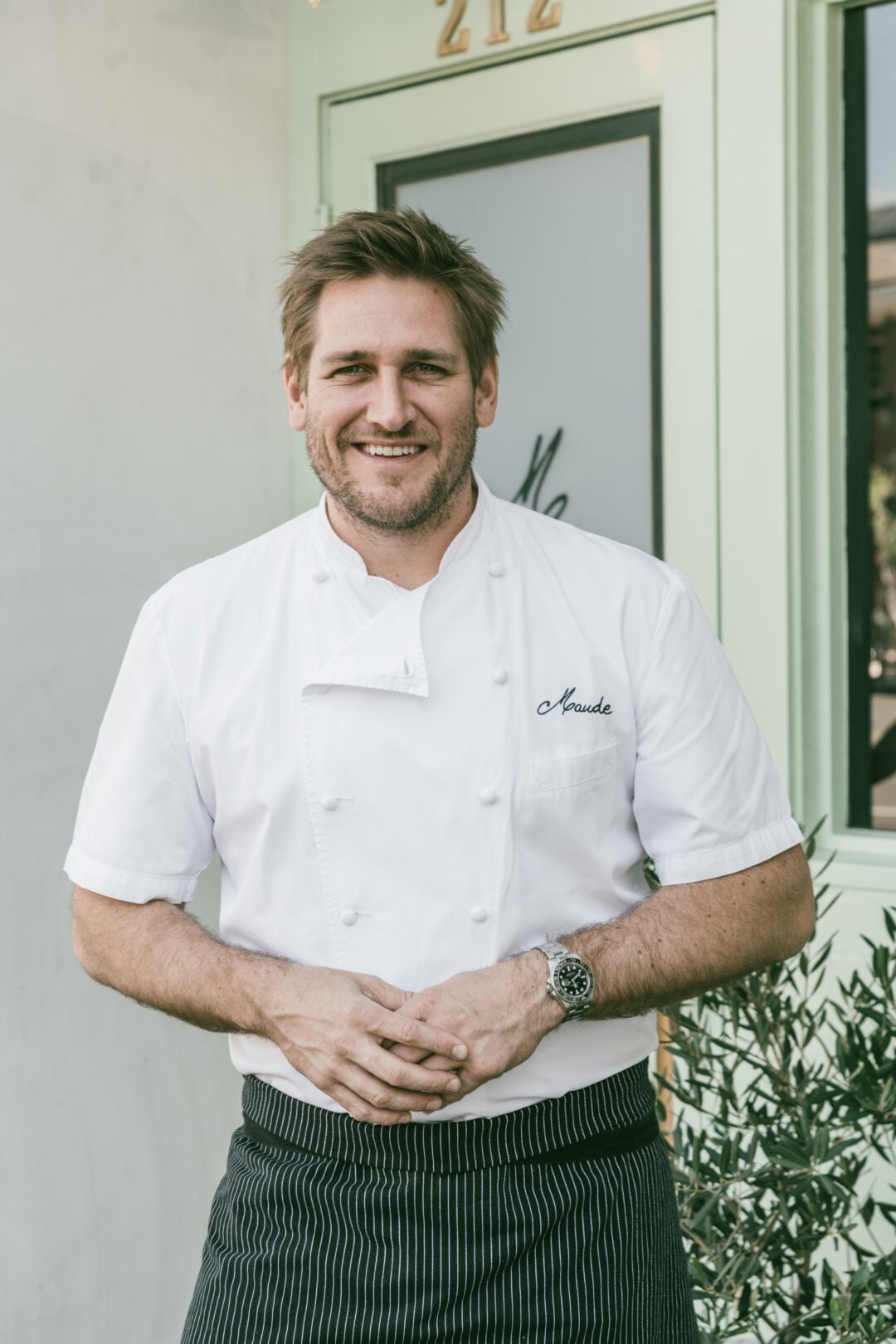 A smiling chef in a white double-breasted chef’s jacket and striped apron stands outside a restaurant door, hands clasped in front of him, with greenery beside him.