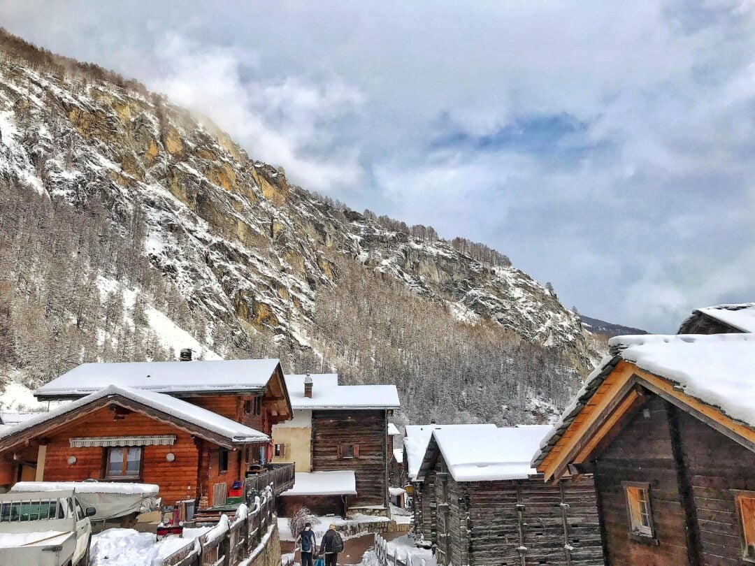 A snowy mountain village with wooden houses, snow-covered roofs, and a rugged, tree-lined mountainside in the background. Two people walk along a path between the houses under a cloudy sky.