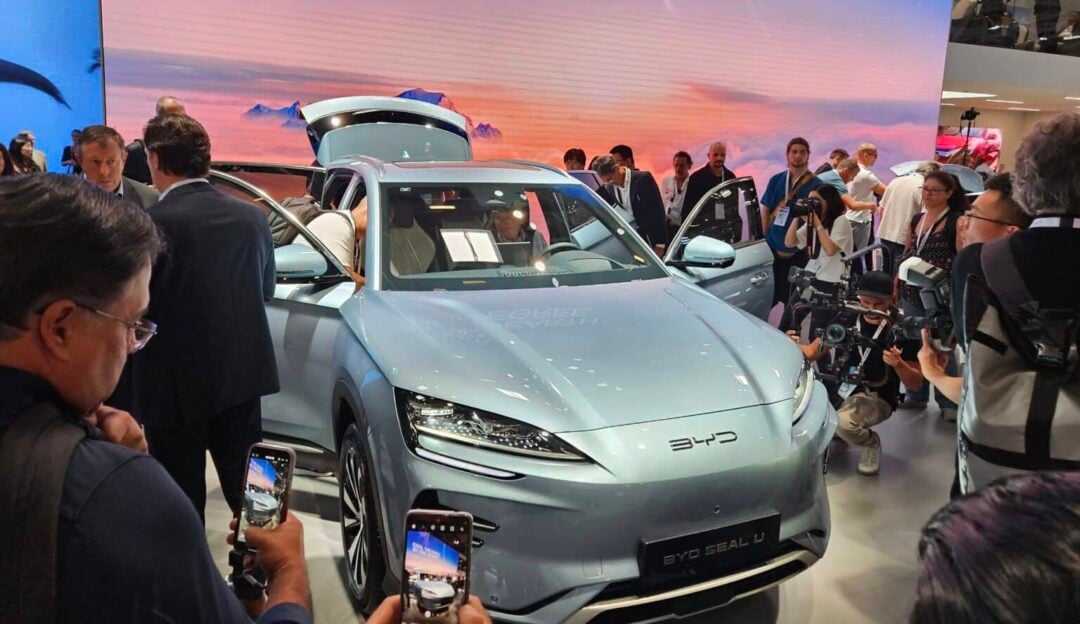 A crowd gathers around a silver BYD Seal U electric car on display at an auto show, with people taking photos and journalists filming the vehicle from multiple angles.