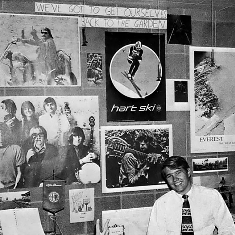 A man in a shirt and tie smiles in an office decorated with posters, including musicians, a skier, Everest, and a quote above: "We've got to get ourselves back to the garden.