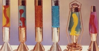 Six colorful lava lamps with metallic bases are displayed in a row against a neutral background. Each lamp contains different colored liquids and wax formations, including glitter and swirling shapes.