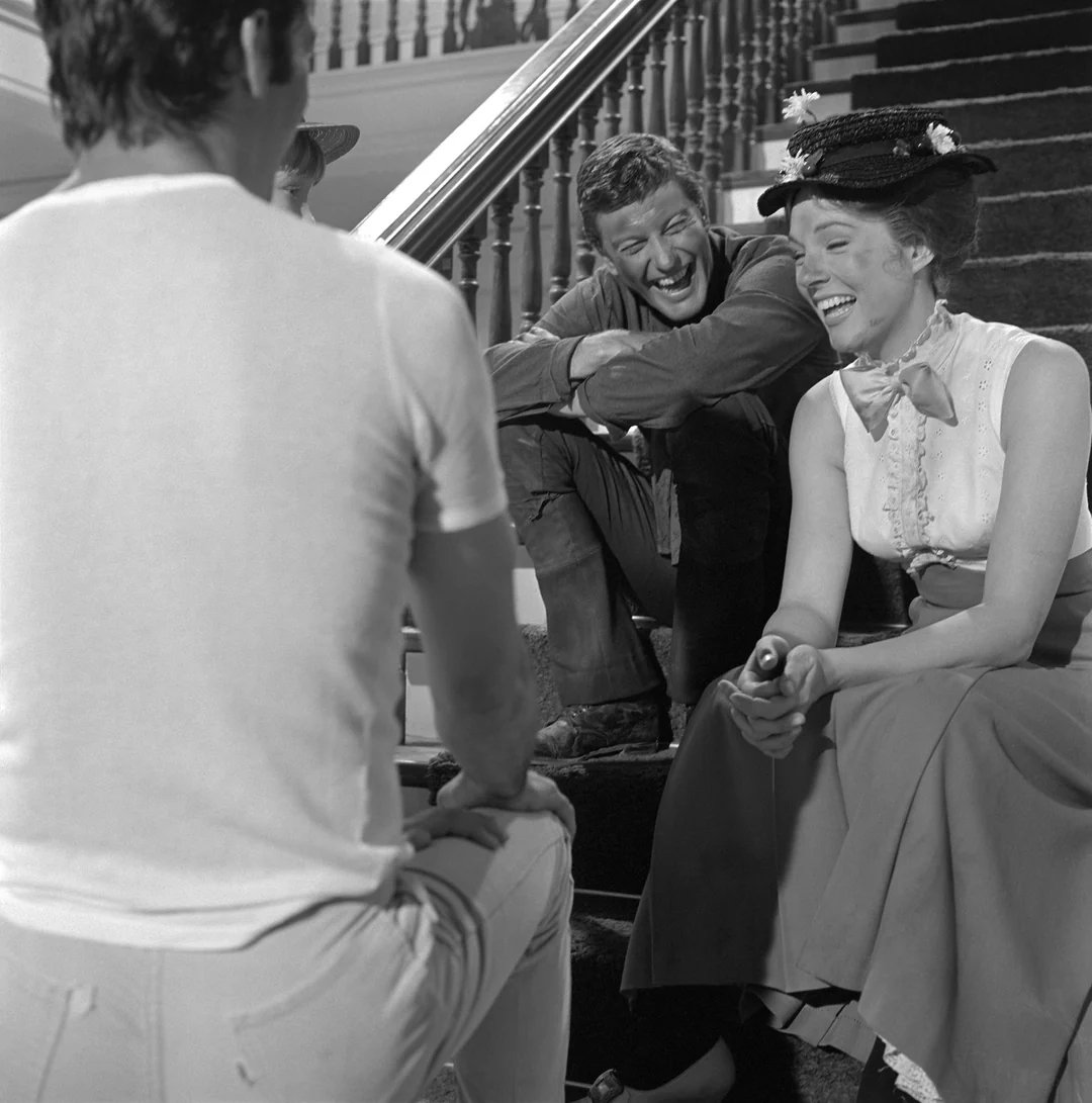 A woman in a period costume and hat laughs while sitting on stairs with two men. One man sits beside her, also smiling, while the other, in a t-shirt, sits with his back to the camera.