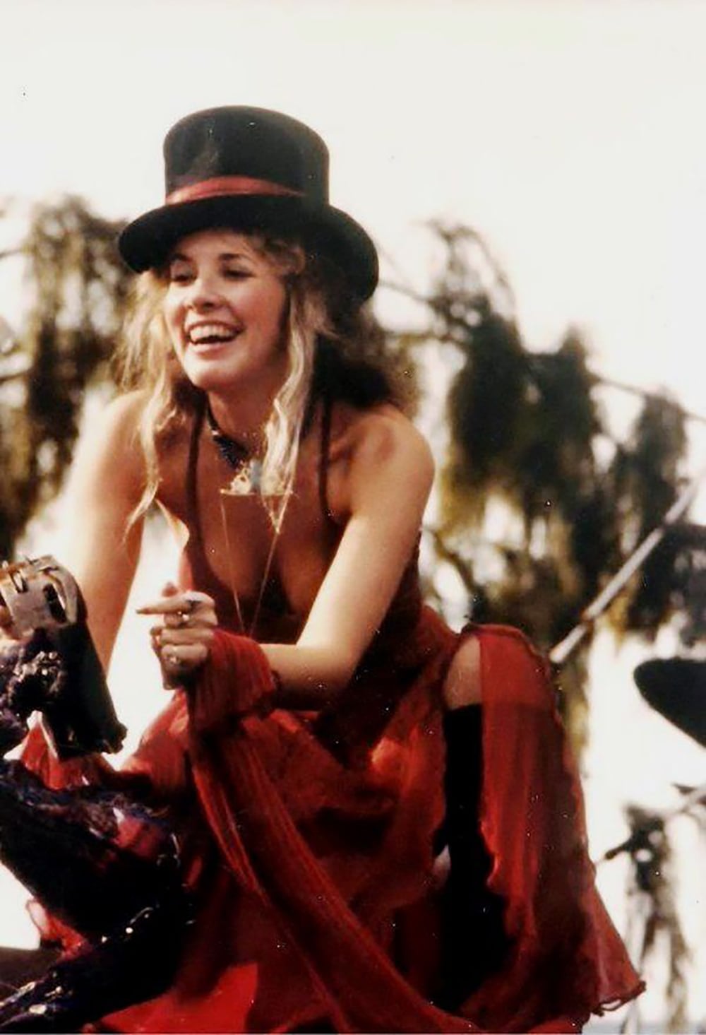 A woman with long blonde hair wearing a black top hat and a flowing red dress smiles while performing outdoors, with trees and stage equipment visible in the background.