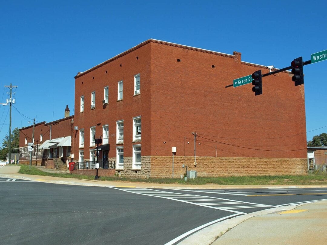 A large red brick building sits on the corner of Green Street and Washington Street under a clear blue sky. The street is freshly paved with marked crosswalks and traffic lights visible.