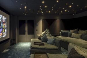 A cozy home theater room features plush couches, a large screen displaying movie covers, soft lighting, and a ceiling with small, star-like lights. The atmosphere is inviting and modern, perfect for watching movies.