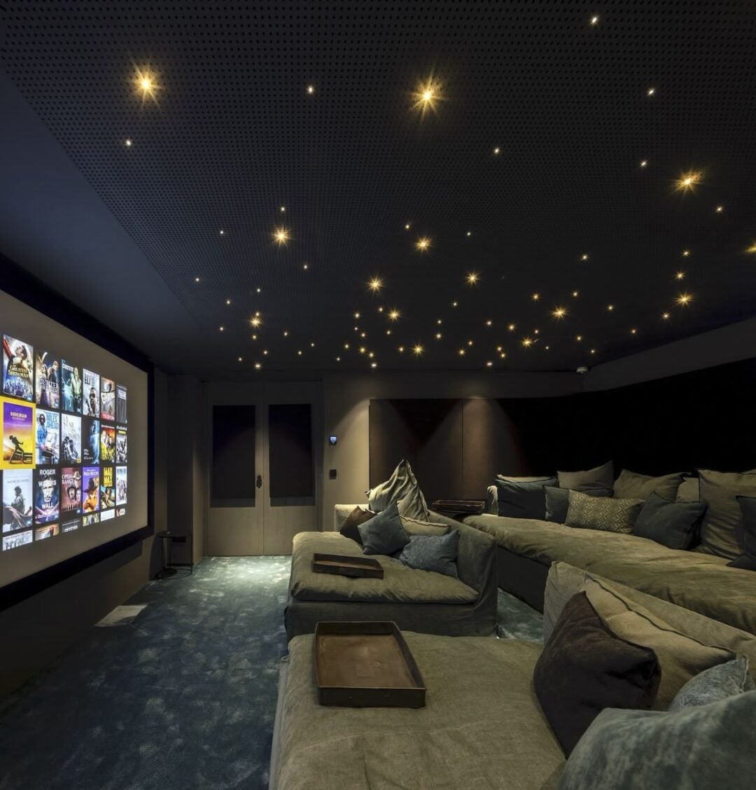 A modern home theater room with tiered, cushioned seating, a large screen displaying movie options, and a ceiling with small star-like lights. The room has a cozy, dimly lit ambiance.