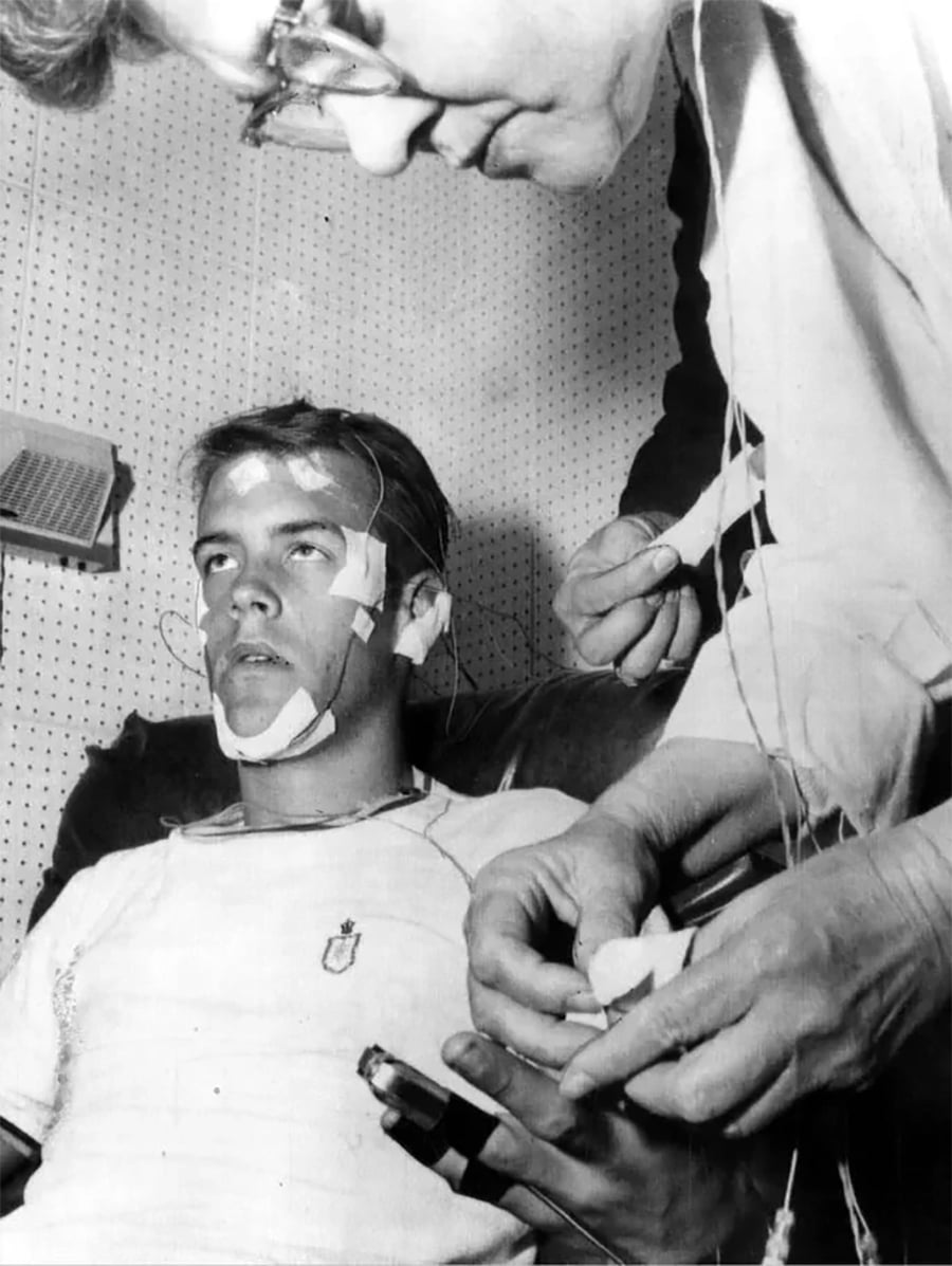 A young man sits with electrodes attached to his face and scalp, likely for a sleep study, while another person adjusts wires and monitors his condition in a clinical setting.