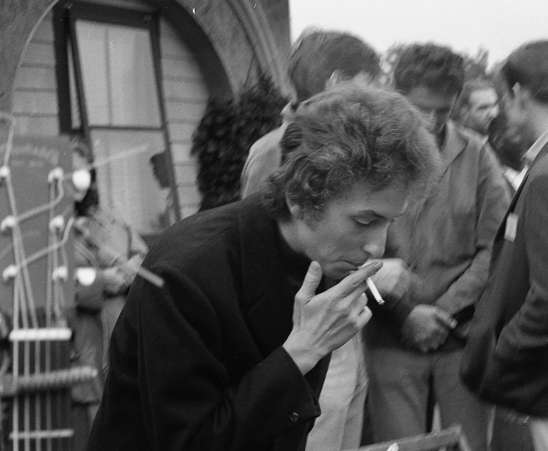 A man with curly hair and a dark jacket leans forward to light a cigarette, surrounded by a small group of people outdoors. The photo is black and white and appears candid, with blurred figures in the background.