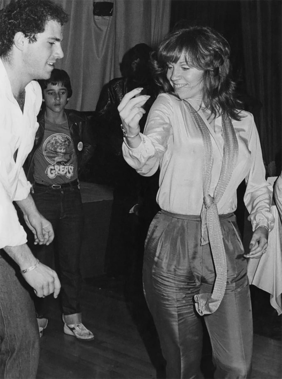 A woman in a shiny blouse and pants dances and smiles at a man in a white shirt. Another young woman in a "Grease" T-shirt and leather jacket stands in the background, watching. The setting appears to be a casual indoor event.
