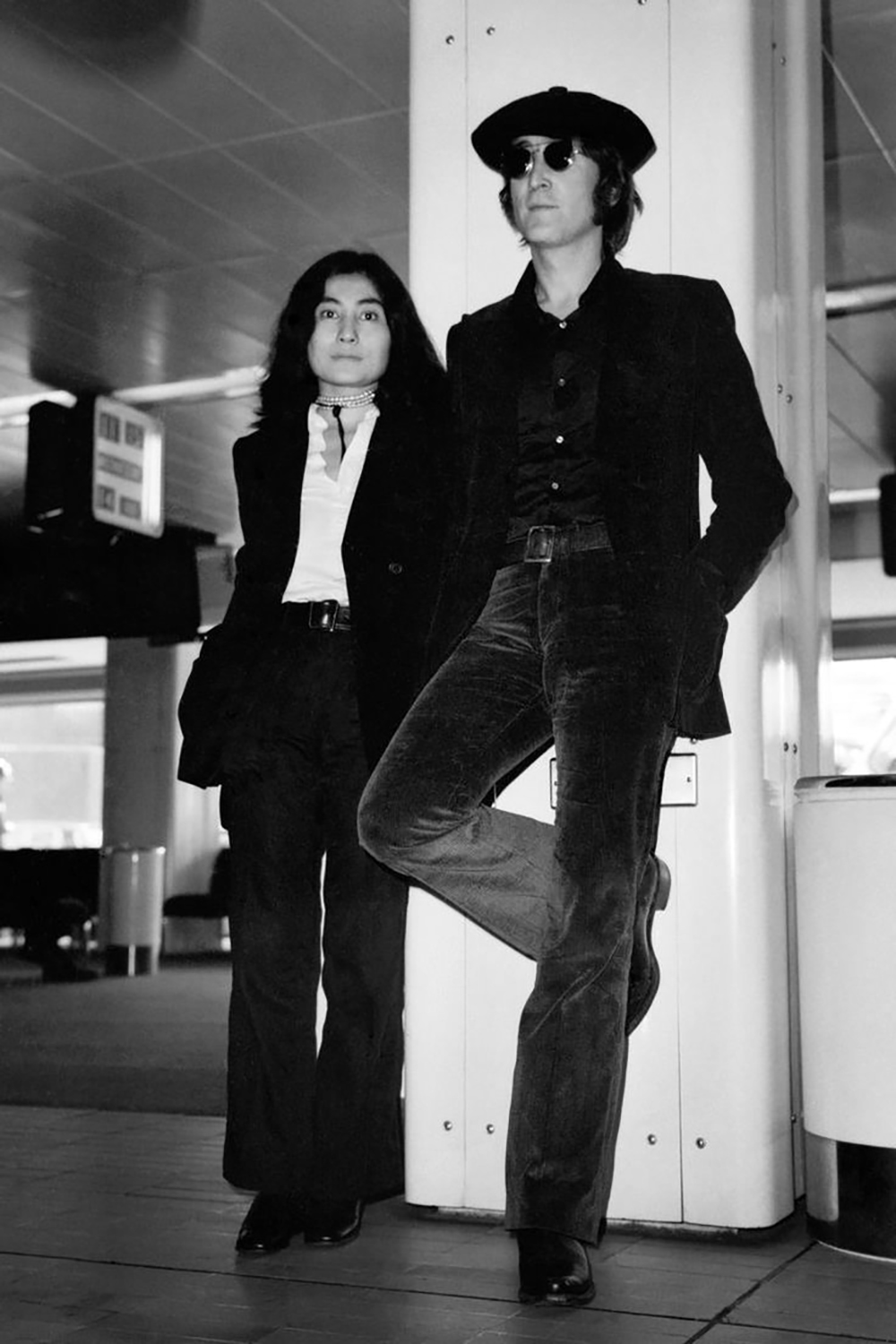 A woman and a man dressed in dark suits stand and lean against a column in an indoor public space. The man wears sunglasses and a hat, while the woman has long dark hair. Both look relaxed.