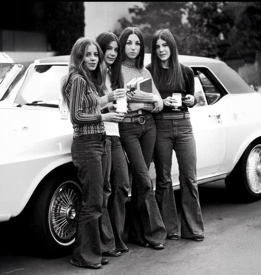 Four young women in 1970s clothing stand together in front of a classic white car, holding drinks and books, smiling at the camera. The image is black and white.
