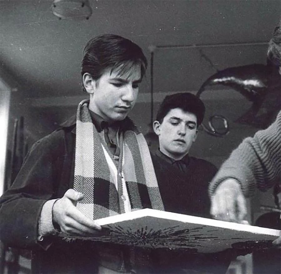 A black-and-white photo of two young men indoors. One holds a rectangular canvas and wears a plaid scarf. The other stands behind him. Another person's arm is visible, pointing at the canvas. A dolphin decoration hangs in the background.