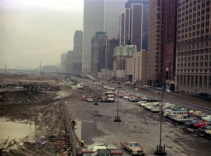 A 1970s cityscape shows a foggy day with parked cars in a nearly empty lot beside tall buildings and a muddy, rubble-strewn area, suggesting ongoing construction or demolition nearby.