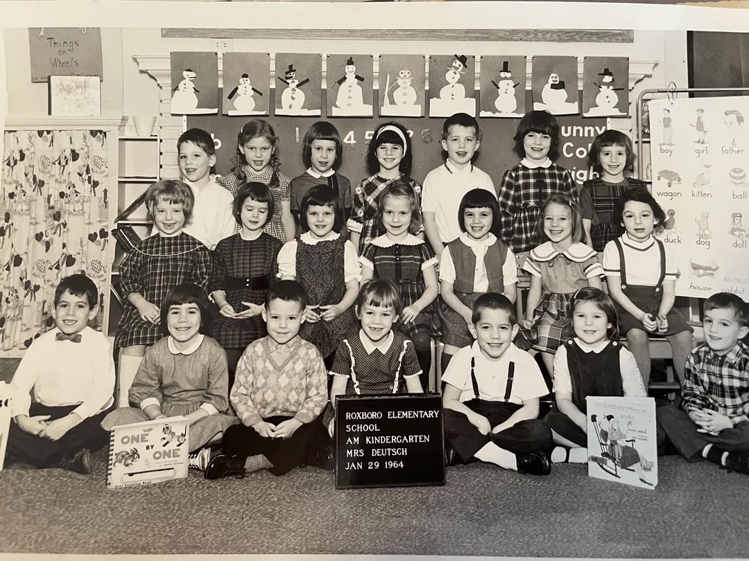 A black-and-white photo of a 1960s kindergarten class. Nineteen children sit and stand in three rows, smiling at the camera. A teacher stands at the back. A sign in front reads "Roxboro Elementary School, Jan 29, 1964.