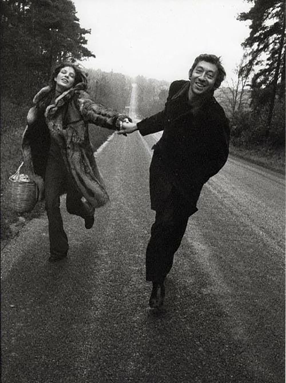 A woman and a man joyfully run hand-in-hand down an empty rural road, smiling. The woman wears a fur coat and carries a basket, while the man is dressed in dark clothing. Trees line both sides of the road. The image is in black and white.