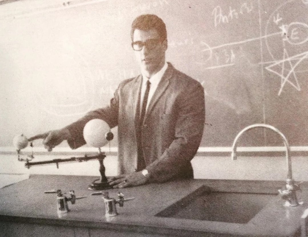 A man in a suit and glasses stands at a lab counter, demonstrating a model of the solar system. He is in front of a chalkboard covered with diagrams and equations.