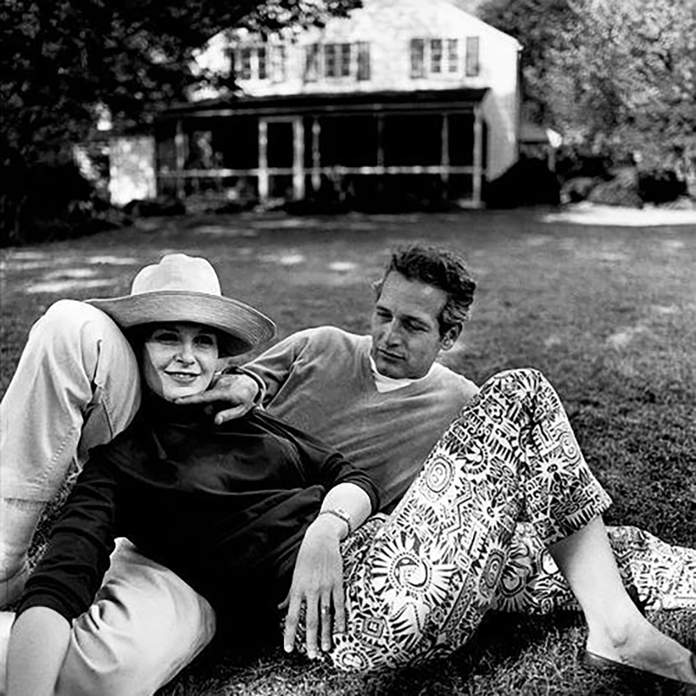 A woman in a wide-brimmed hat and patterned pants relaxes on the grass, leaning against a man in a sweater. They sit together in front of a large house with a porch, surrounded by trees.